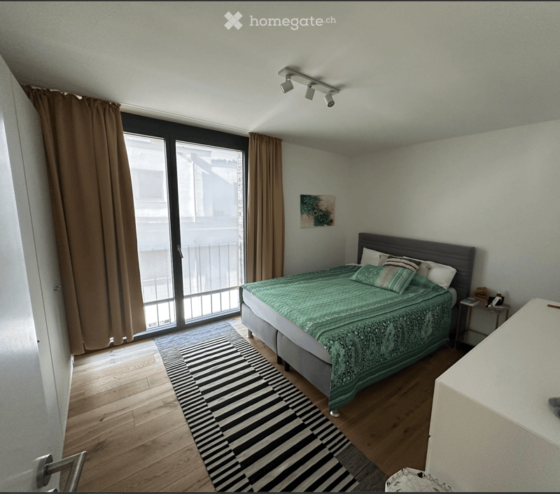 Modern bedroom with wooden flooring, large window with sliding glass doors, bed with green duvet, nightstand, and artwork on the wall.