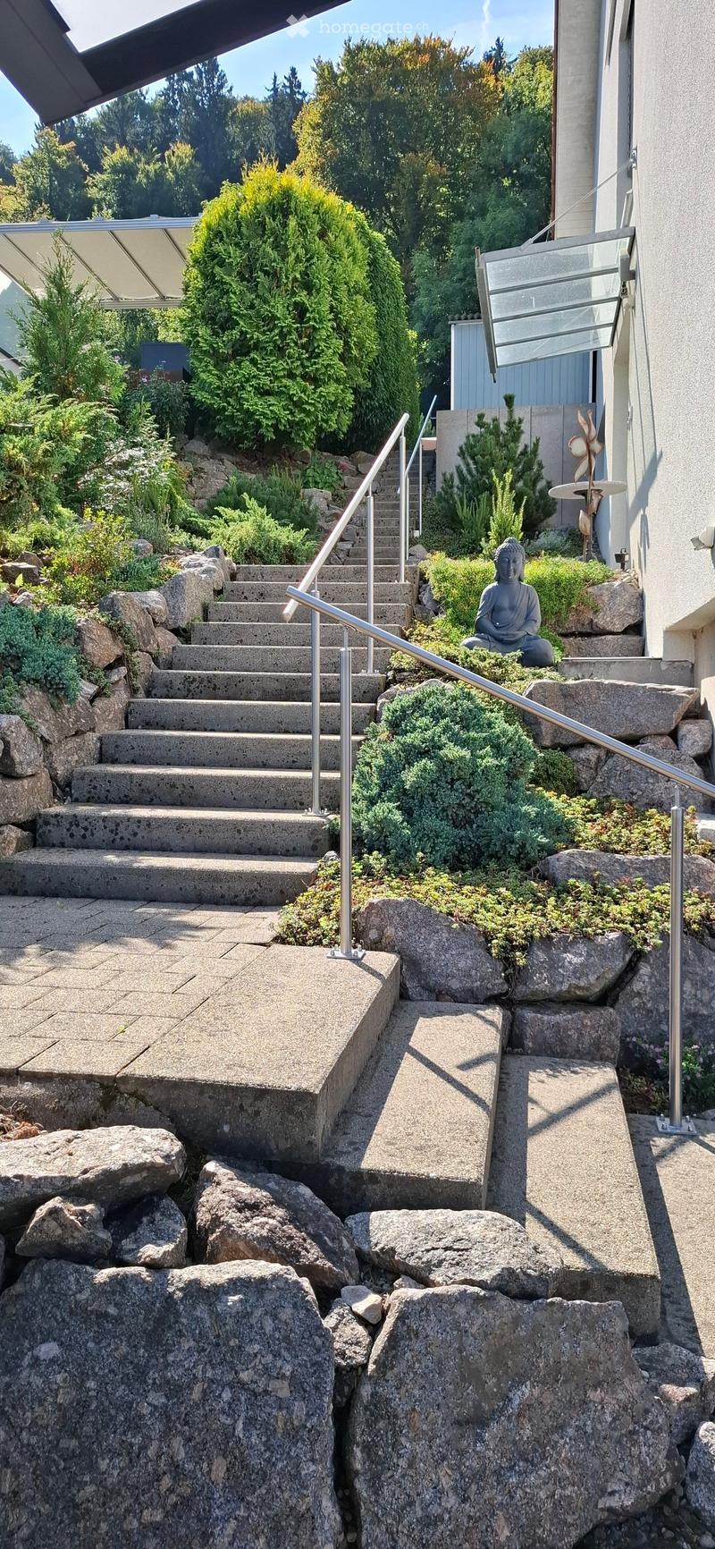 A landscaped garden with a stone staircase, metal handrails, and various plants and rocks.