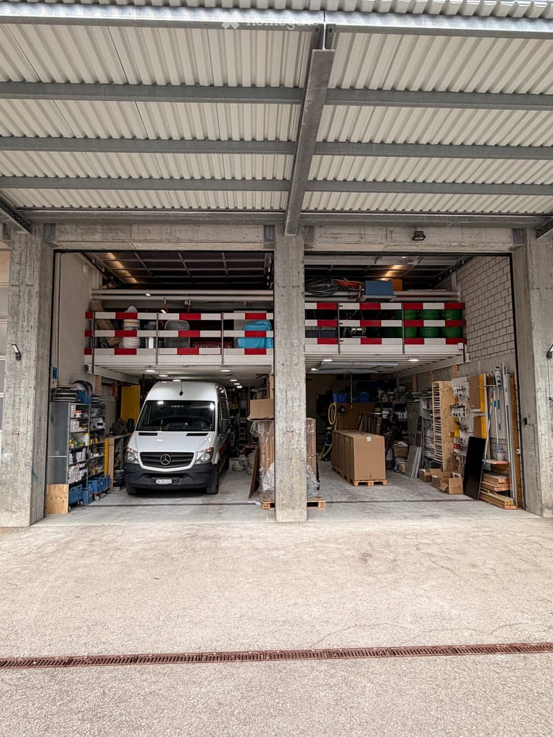 Large, open garage with concrete floors and walls, featuring a parked van, shelves with storage items, and various tools and equipment.