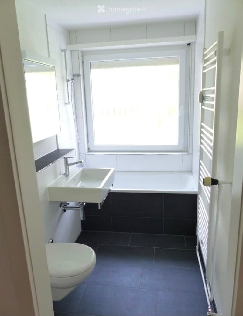 Modern bathroom with white tiles, black floor tiles, a large window, a sink, a toilet, a bathtub, and a radiator.