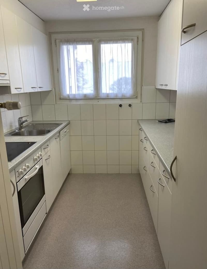 A small, unfurnished kitchen with white cabinets, a sink, an oven, and a window with blinds.