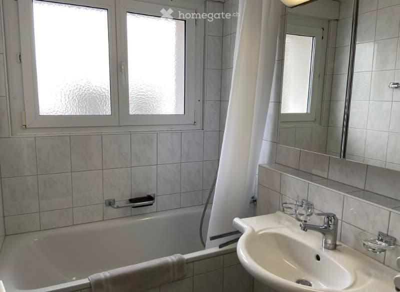 Bathroom with a bathtub, sink, mirror, and towel. The room features tiled walls and a window with frosted glass.