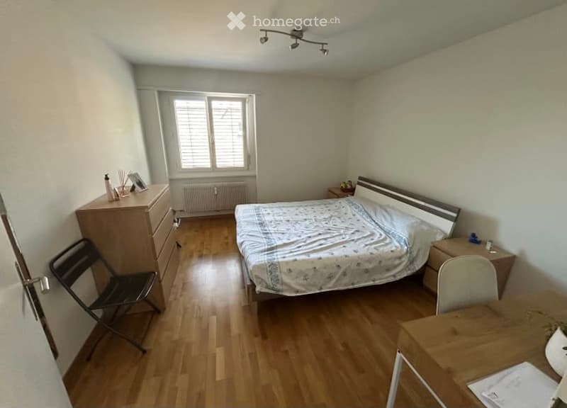 A furnished bedroom with a bed, nightstand, chair, desk, and drawer. The room features wooden flooring and a window with blinds.