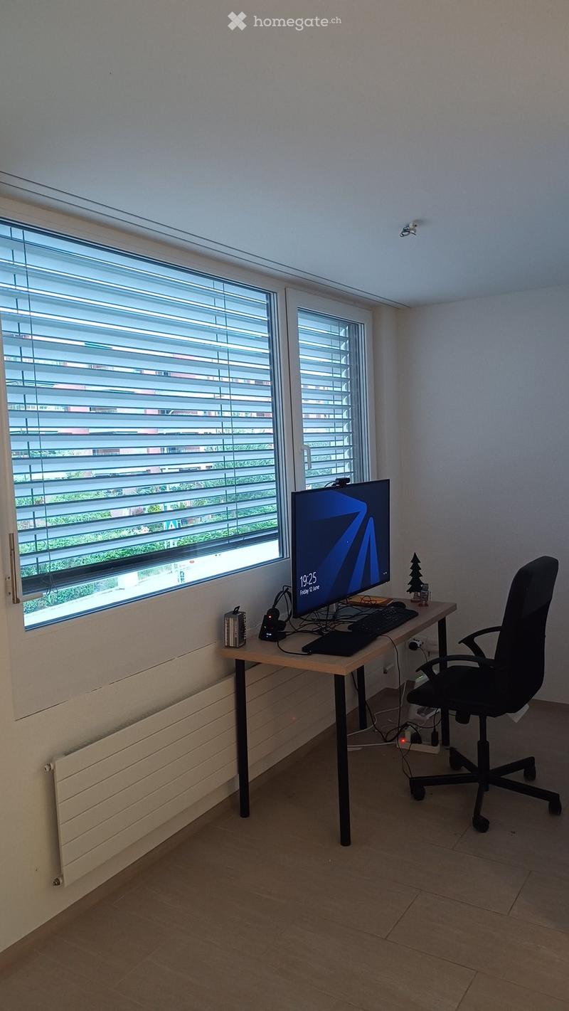 A modern, well-lit room with a desk setup, a computer monitor displaying a digital clock, a comfortable office chair, and a small decorative tree. The room features large windows with blinds and a radiator below them.