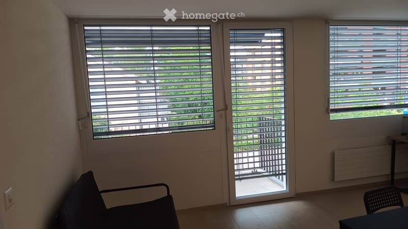 Empty room with wooden flooring, large windows with blinds, and a sliding glass door leading to a balcony.