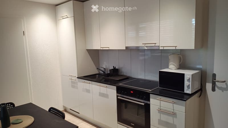 Modern kitchen with white cabinets, stainless steel appliances, and a black countertop. Includes a sink, oven, microwave, teapot, and a small dining area with a table and chairs.