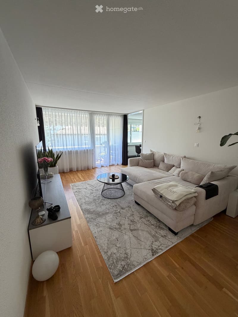 Modern living room with a sectional sofa, coffee table, area rug, and decorative items including a large white ball and a vase with flowers. Large windows with sheer curtains let in natural light.