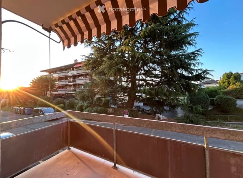 Balcony with striped awning, overlooking a tree-lined street and multi-story apartment building, with sunset visible.