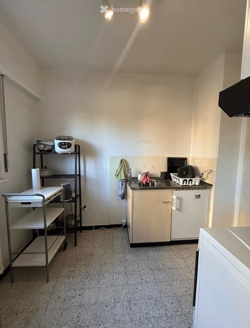 A small kitchen with white cabinets, a sink, a microwave, an oven, a cart, and a refrigerator. The floor is tiled, and there are various kitchen items on the counter and shelves.