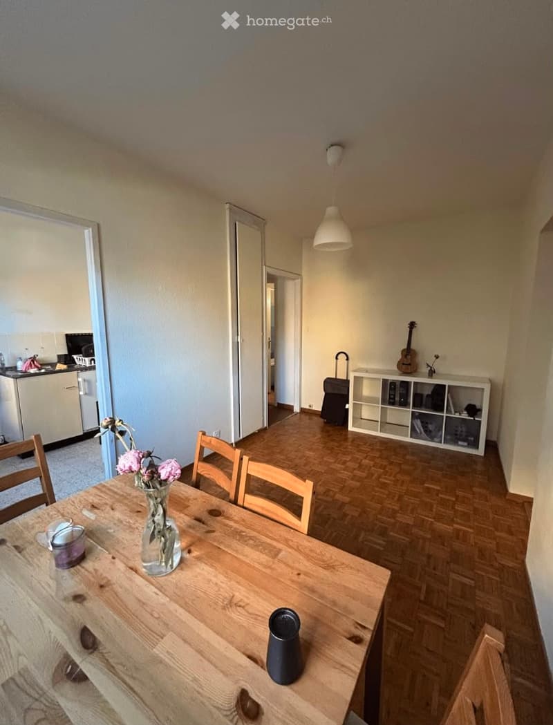A well-lit dining area with a wooden table, chairs, a vase with flowers, and a guitar on a shelf. The room features wooden flooring and a modern aesthetic.