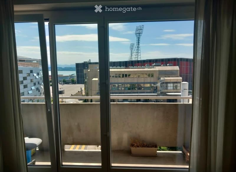 Balcony with a view of a cityscape, including a stadium and surrounding buildings. The balcony has a metal railing and a small potted plant.