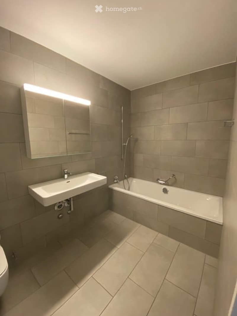 Modern bathroom with a white sink, bathtub, and large mirror with built-in lighting. Grey tiled walls and floor.