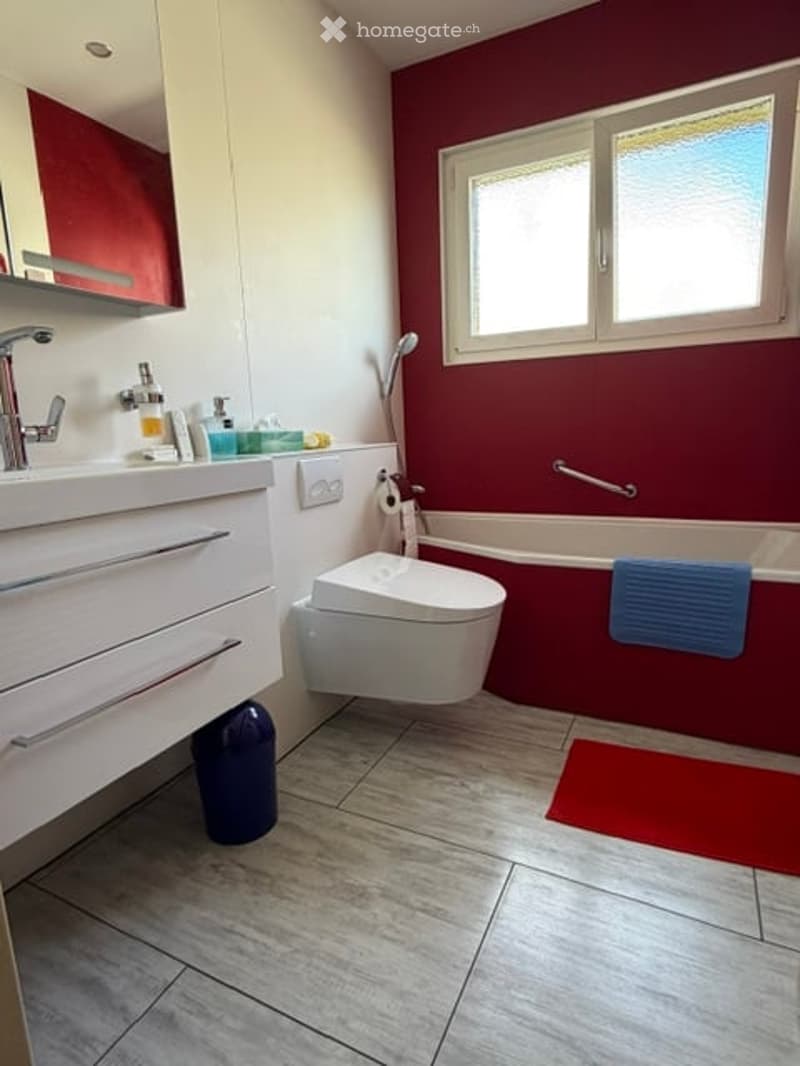 Modern bathroom with red accent wall, white fixtures, and grey tiled floor. Features a sink with a modern faucet, a toilet, a bathtub with a blue towel, and a shower in the corner.