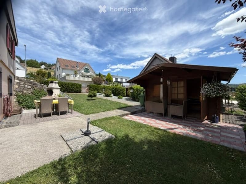 Well-maintained garden with a patio area, outdoor seating, and a small wooden structure, surrounded by greenery and neighboring houses.