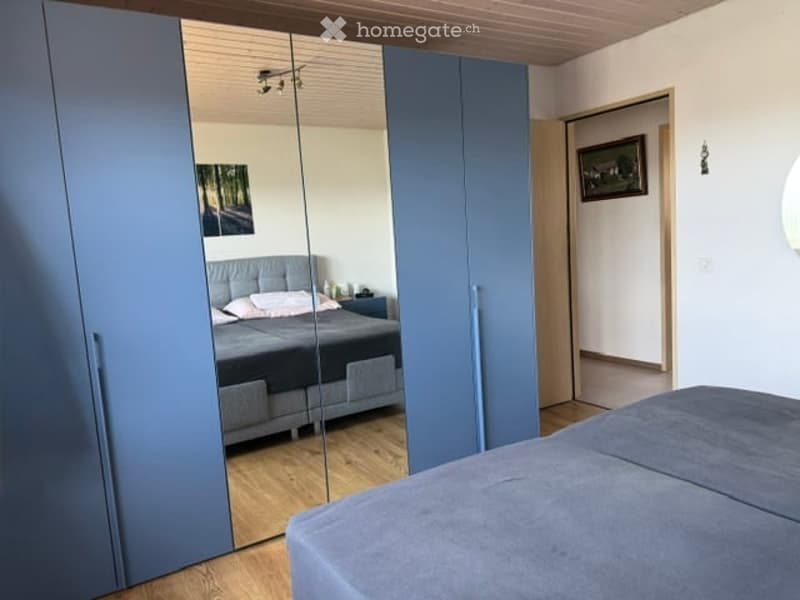 Modern bedroom with a grey bed, nightstand, large mirrored wardrobe, and wooden flooring.