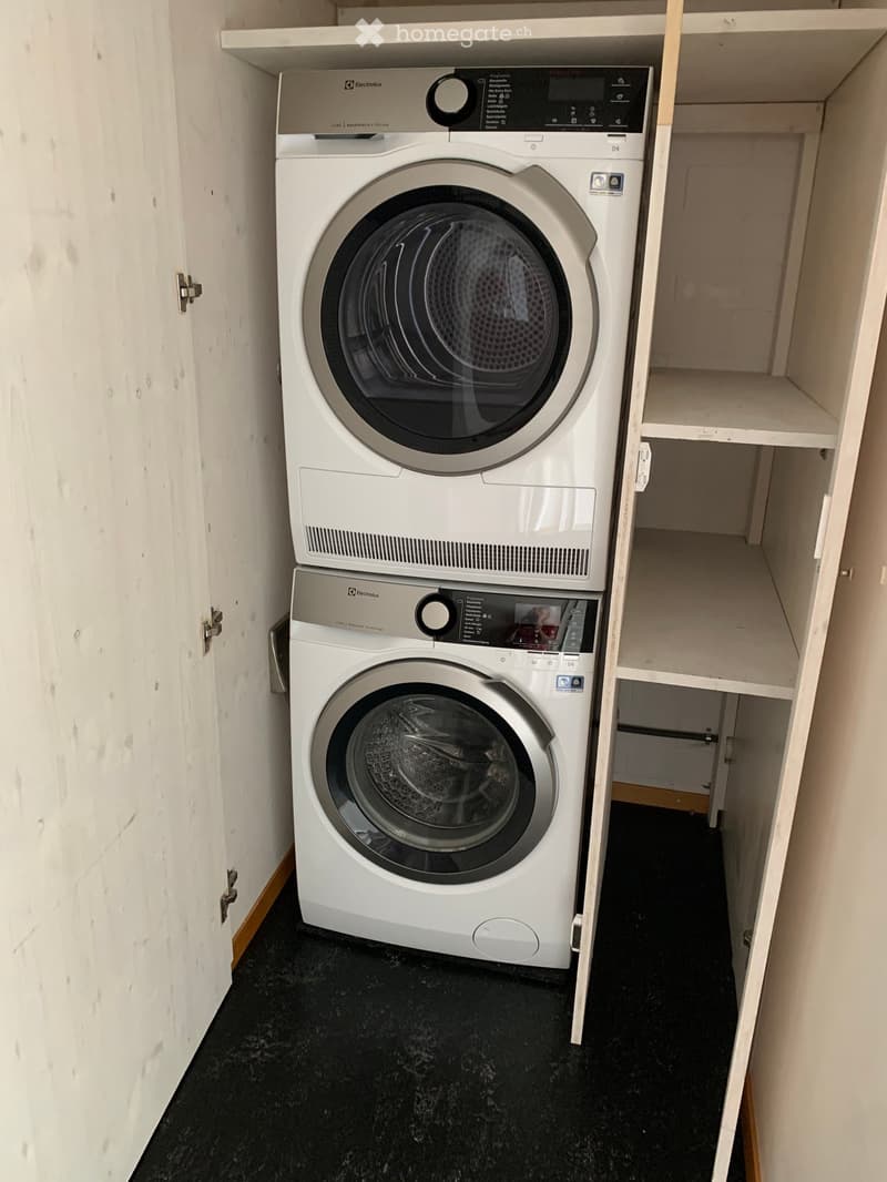 A compact bathroom with a white washing machine and a white dishwasher placed side by side, with built-in white shelving units on the right side.