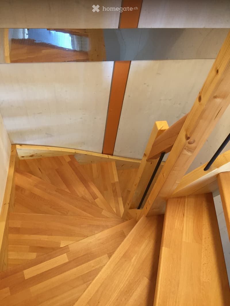 Spiral staircase with wooden steps and handrails, leading upwards, with a modern interior design featuring light-colored walls and a glass window at the top.