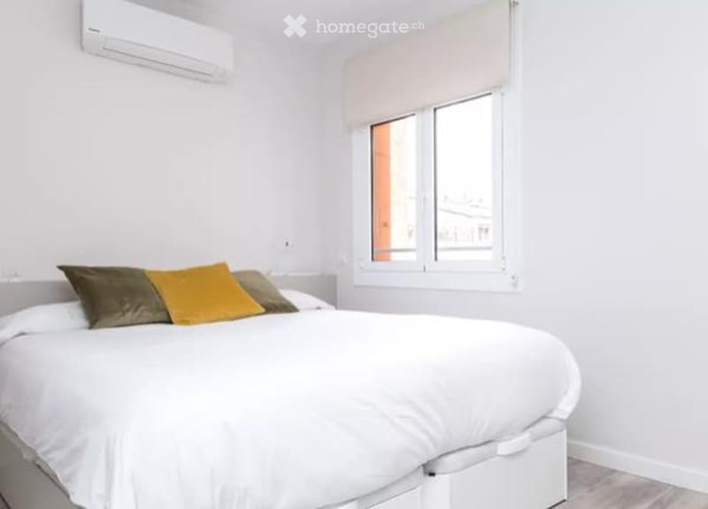 Modern bedroom with a large bed, multiple pillows, a window with blinds, and an air conditioner mounted on the wall.