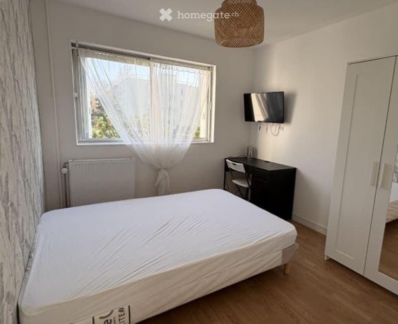 Cozy bedroom with a single bed, a desk with a chair, a wall-mounted TV, a large window with white curtains, and a wardrobe with a mirror.