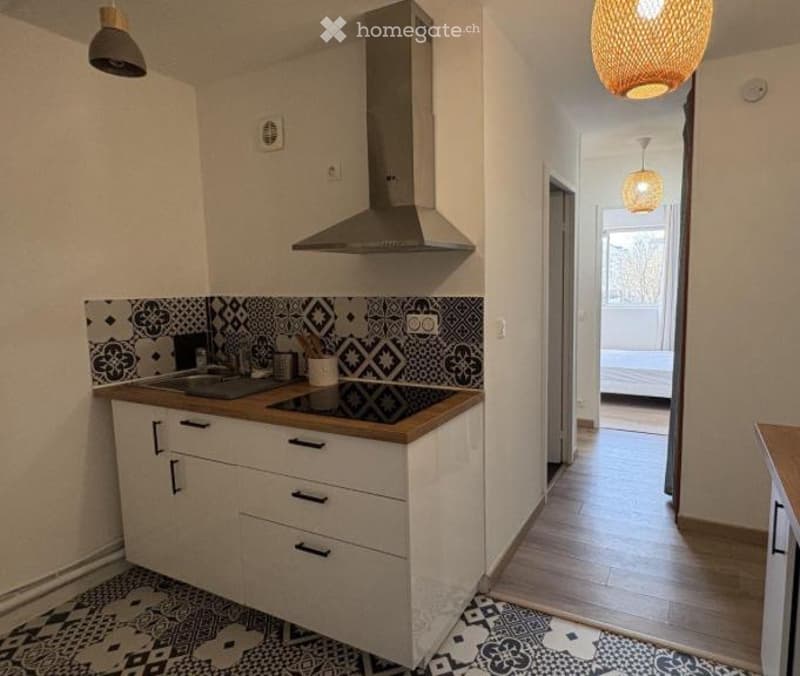 Modern kitchen with white cabinets, induction stove, and a range hood. The floor is wooden, and the walls are painted white. A hallway leads to a bedroom with a visible bed.