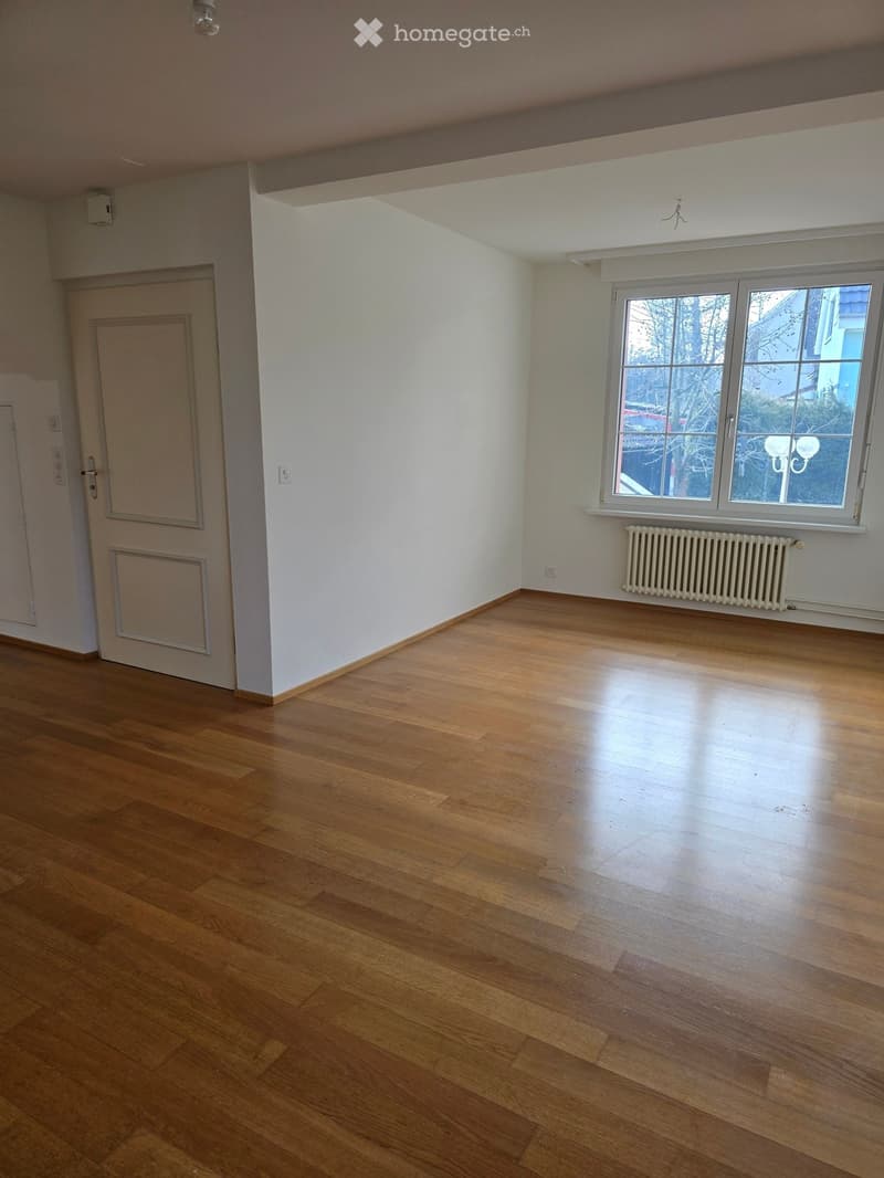 Empty room with wooden flooring, white walls, a window with a radiator below, and a door on the left side.