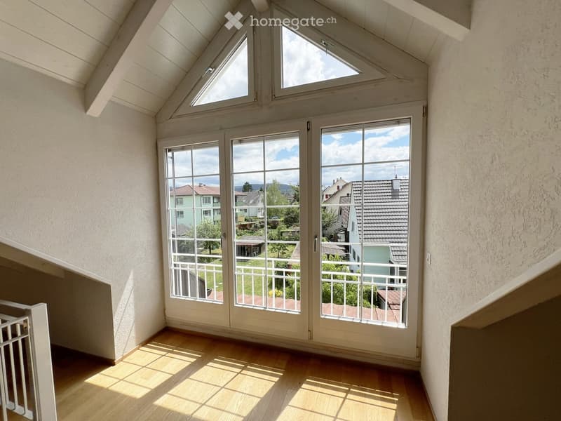 Bright, empty room with wooden flooring, large windows, and a view of a garden and neighboring houses.