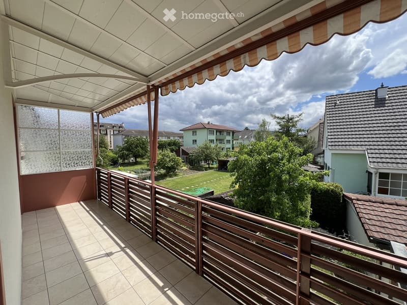 Balcony with tiled floor, wooden railing, and striped awning, overlooking a green garden and neighboring houses.
