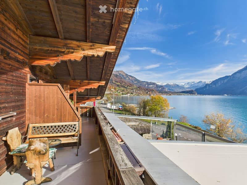 Wooden balcony with a scenic view of a lake and mountains, featuring a bench, chair, and small table.