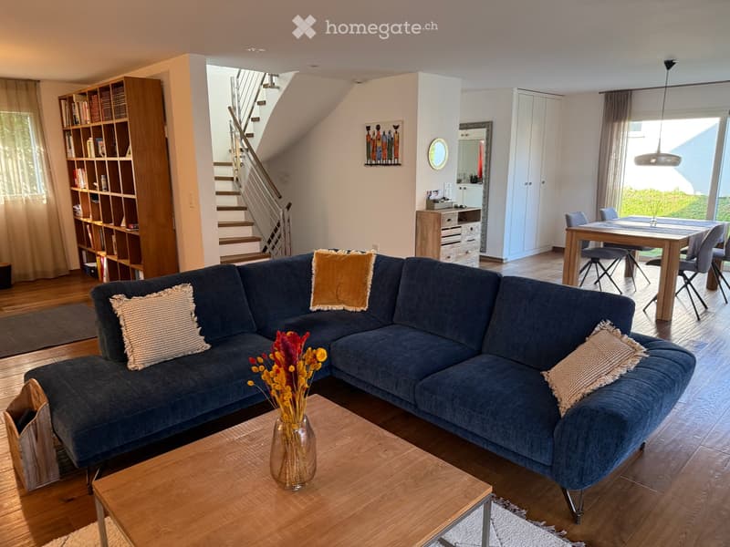 Modern living room with a blue sectional sofa, wooden coffee table, vase with flowers, bookshelf, dining area with table and chairs, and a staircase leading to the upper level.