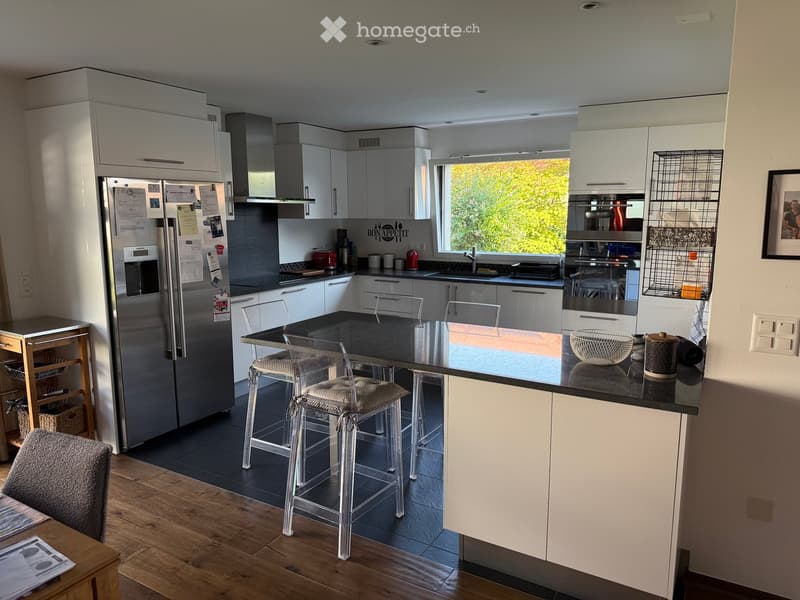 Modern kitchen with white cabinets, stainless steel appliances, island with bar stools, and a large window overlooking greenery.