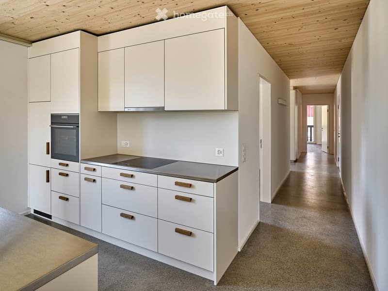 Modern hallway with white walls, grey carpeted floor, and a kitchen area visible on the left side with white cabinets, wooden handles, and a built-in oven.