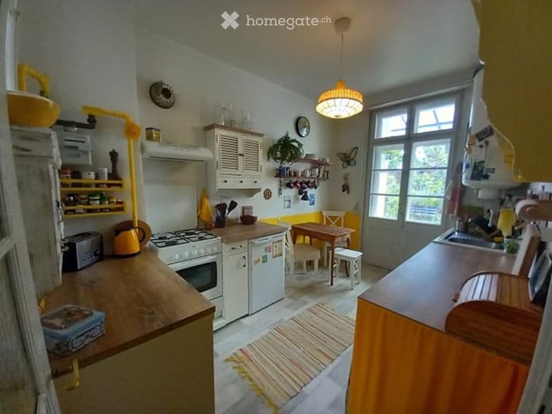 Kitchen. Bright kitchen with white cabinets, a central island, and a small dining area. Appliances include a refrigerator, oven, and stove. A small table with chairs and a stool is present, along with a colorful rug on the floor.