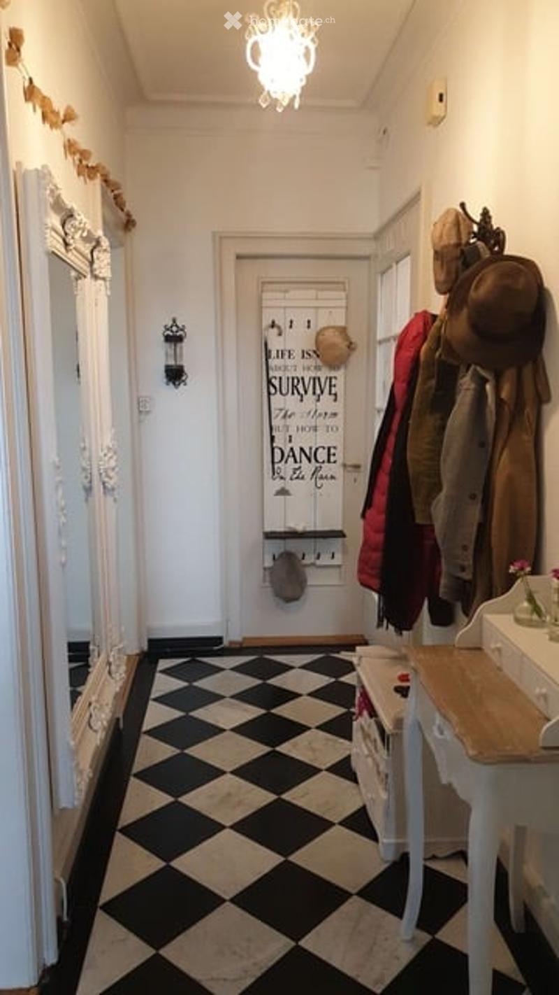 Corridor / Hallway. Hallway with black and white checkered floor, white walls, coat rack with jackets, small table with vase, and decorative wall sign.
