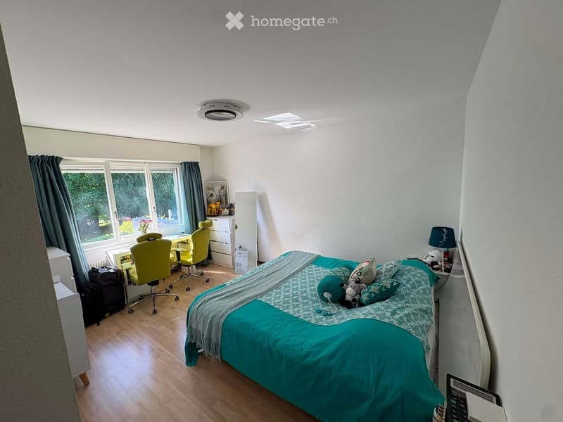 Bright bedroom with a large bed, green bedding, a desk area with chairs, a lamp, and a mirror. Large windows with curtains provide natural light.