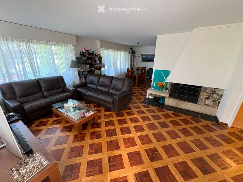Spacious living room with dark leather sofa, glass coffee table, fireplace, dining area with table and chairs, large windows with curtains, and wooden flooring.
