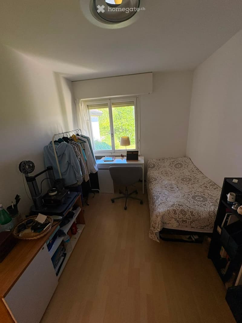 Cozy bedroom with wooden flooring, a bed with patterned bedspread, a desk with a chair, a wardrobe, and a shelf with various items.
