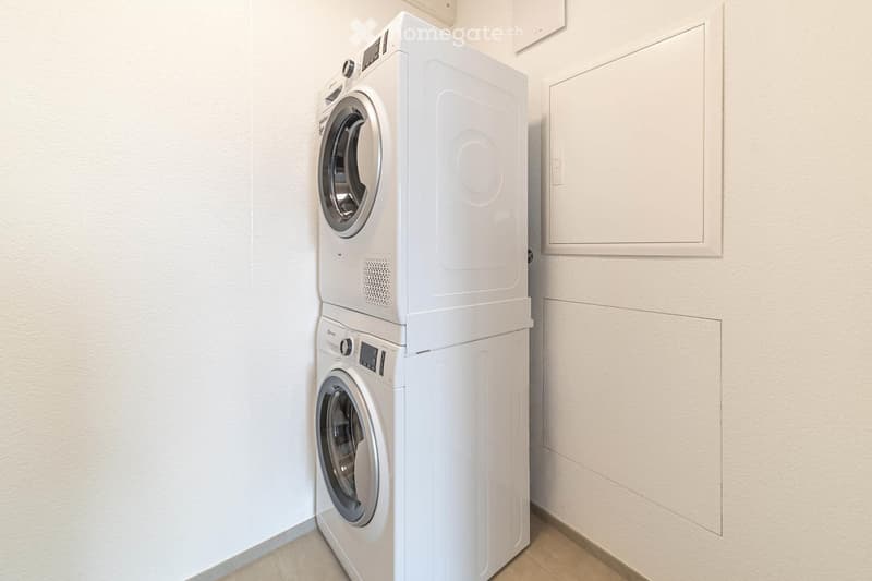 A small, well-lit room with a stacked washer and dryer unit against a white wall. The floor is tiled, and there is a small cabinet to the right.