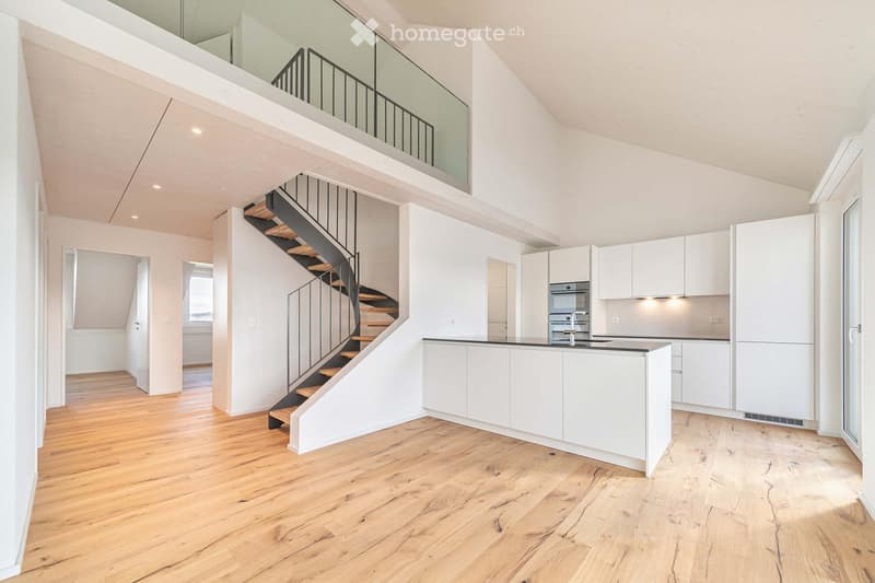 Open-plan space with wooden flooring, white walls, a modern staircase leading to an upper level, and a white kitchen area with cabinets and appliances.