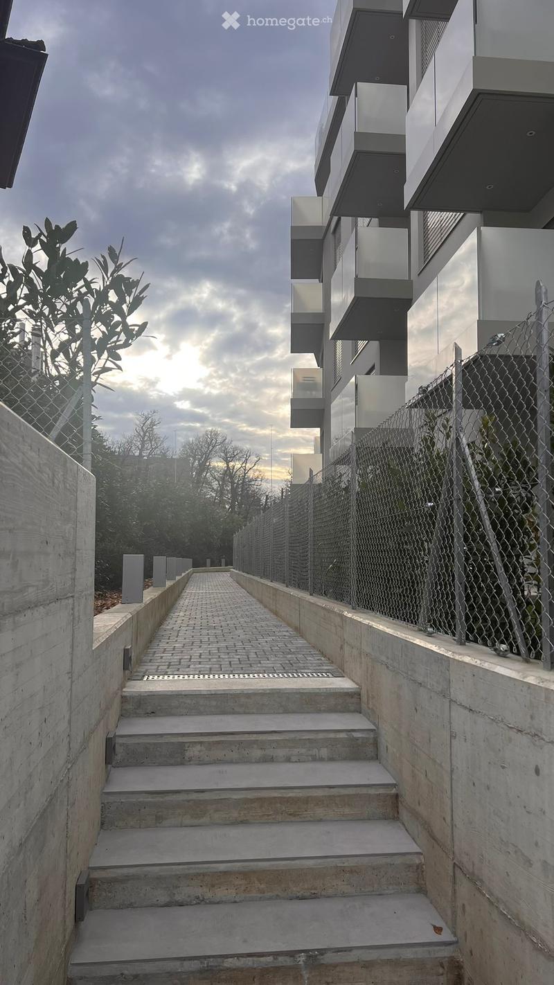 A modern concrete staircase leading up to a contemporary building with glass and metal elements, surrounded by a chain-link fence and greenery.