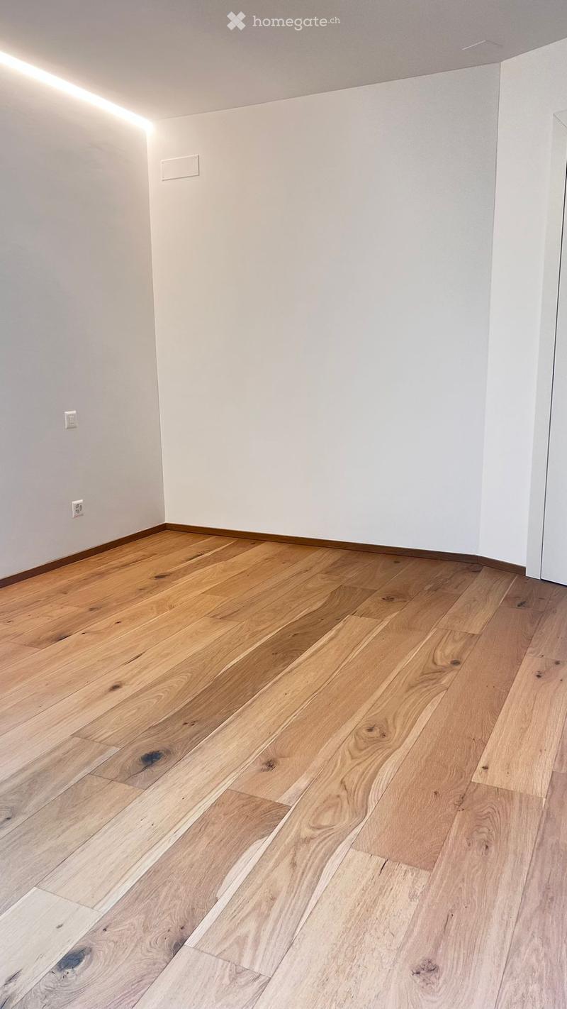 Empty room with light wooden flooring and white walls, featuring a subtle built-in LED light strip along the ceiling edge.