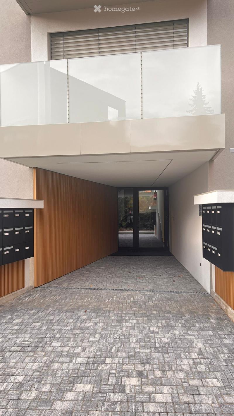 Modern entrance corridor with a paved stone floor, wooden paneling on the walls, and mailboxes on either side. A glass door leads to the interior, with a raised ground floor visible above.