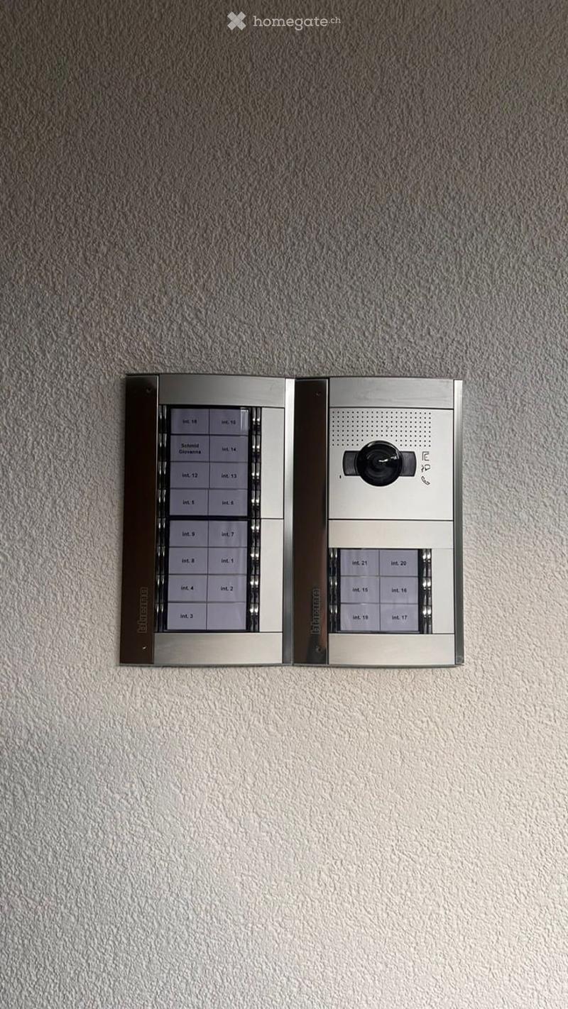 Modern intercom system mounted on a textured wall, featuring multiple apartment numbers and a camera interface.