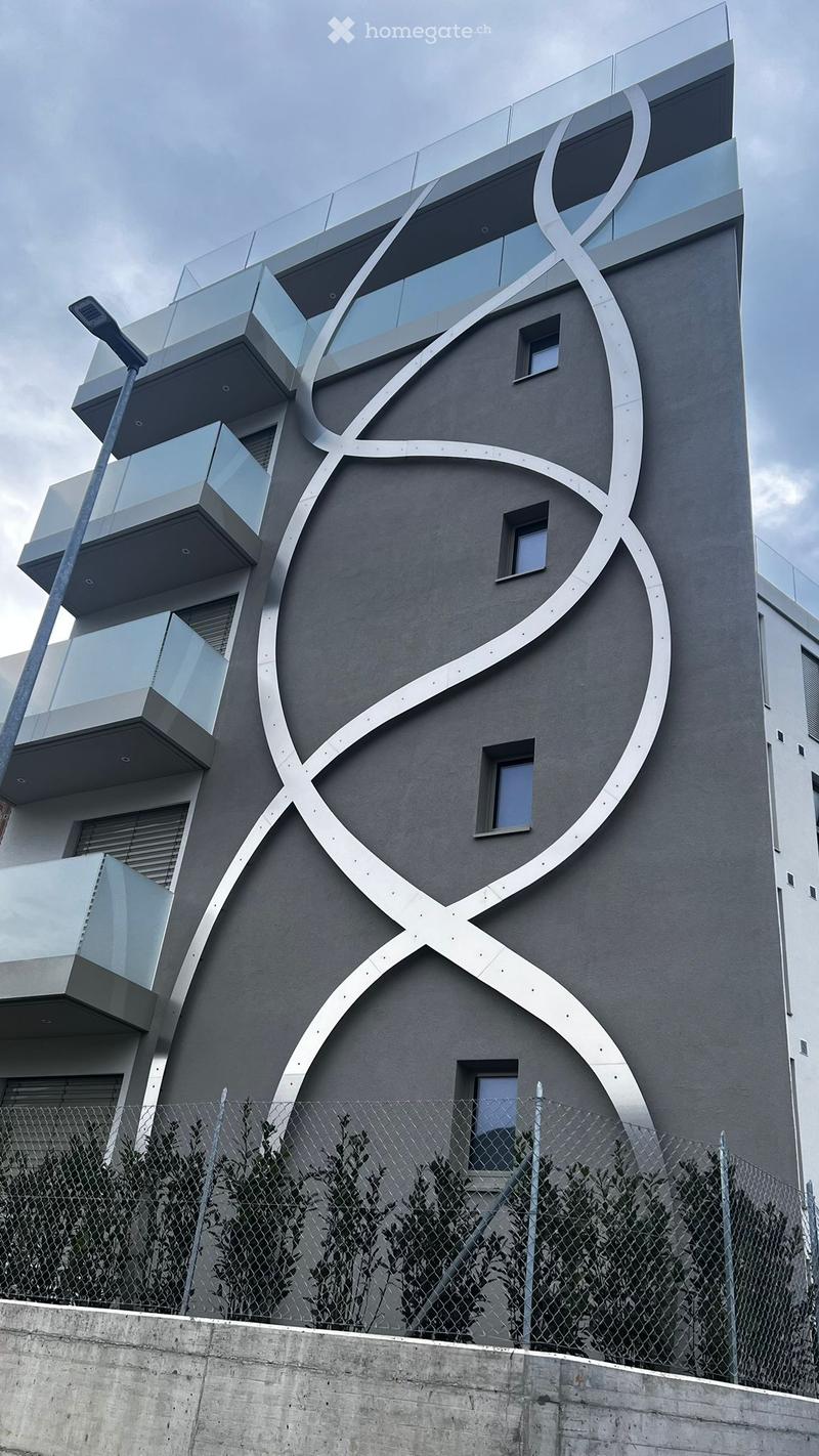 Modern multi-story building with a sleek, grey facade and large, curved metallic decorative elements. Features multiple balconies with glass railings and several windows.