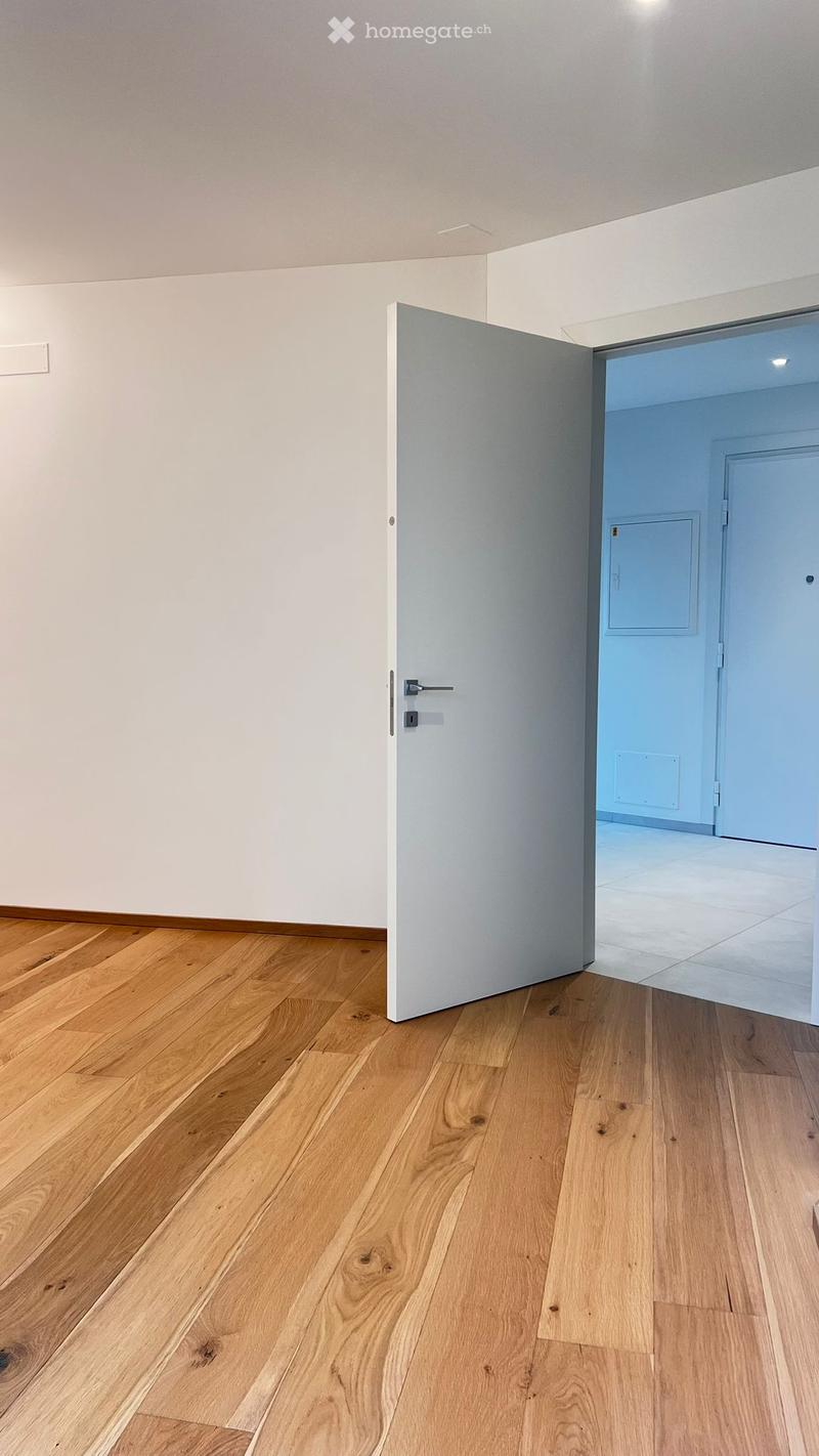 Empty room with light wooden flooring and white walls, featuring an open door leading to another room with light blue walls and tiled flooring.