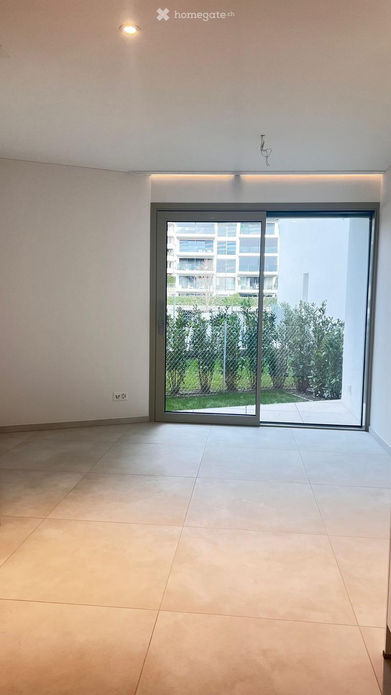 Empty room with tiled floor, large sliding glass door leading to a garden area, modern lighting fixture on the wall, and a power outlet visible on the left wall.