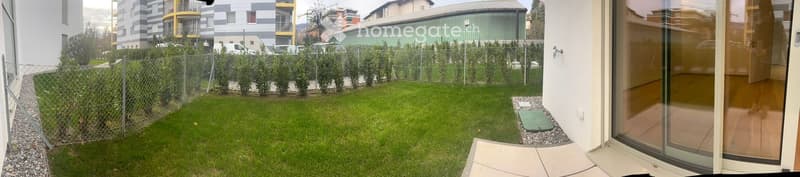 Well-maintained garden with neatly trimmed hedges, a small lawn, and a paved patio area adjacent to a modern building entrance.