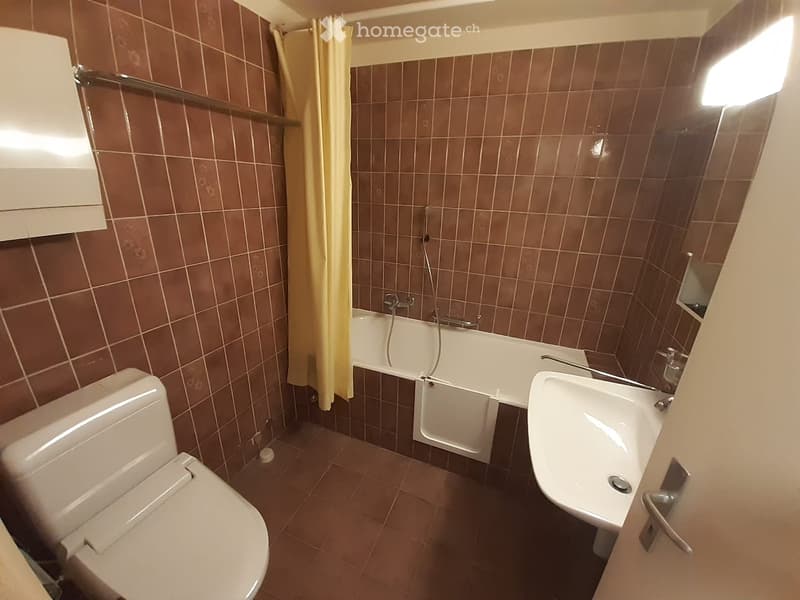 Bathroom with brown tiled walls and floor, featuring a toilet, bathtub with a shower curtain, and a sink.