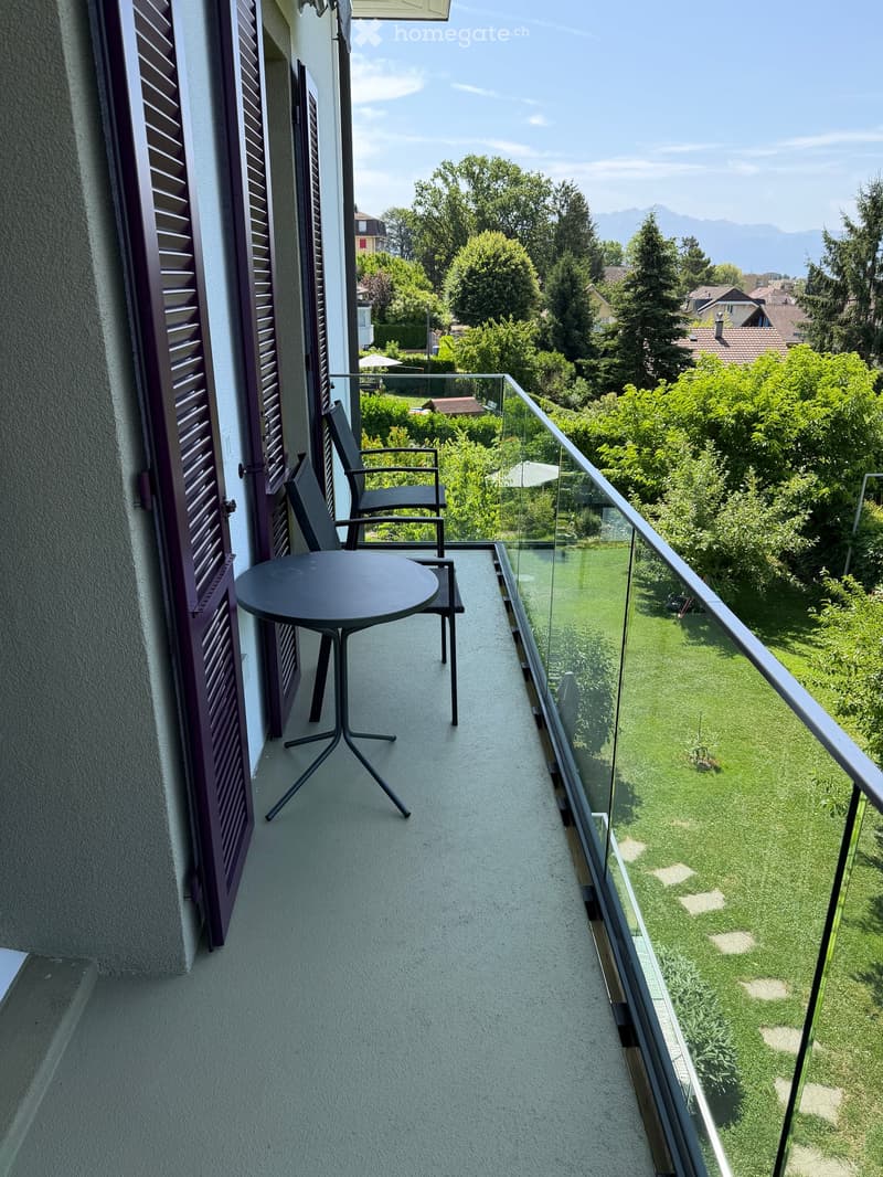 Balcony with a round table and two chairs, overlooking a green lawn and trees.