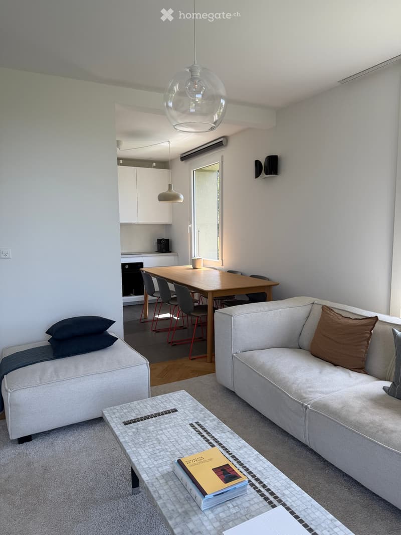 Modern living room with a grey sectional sofa, coffee table with books, dining table with chairs, and an open kitchen area with white cabinets.