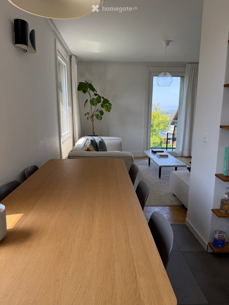 Bright living room with a sofa, coffee table, dining table, chairs, pendant lights, and a view through large windows.